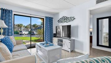 Ein modernes Wohnzimmer mit Sofa, Couchtisch und Fernseher. Eine Glastür öffnet sich zum Balkon mit Blick auf die Straße und geparkte Autos.