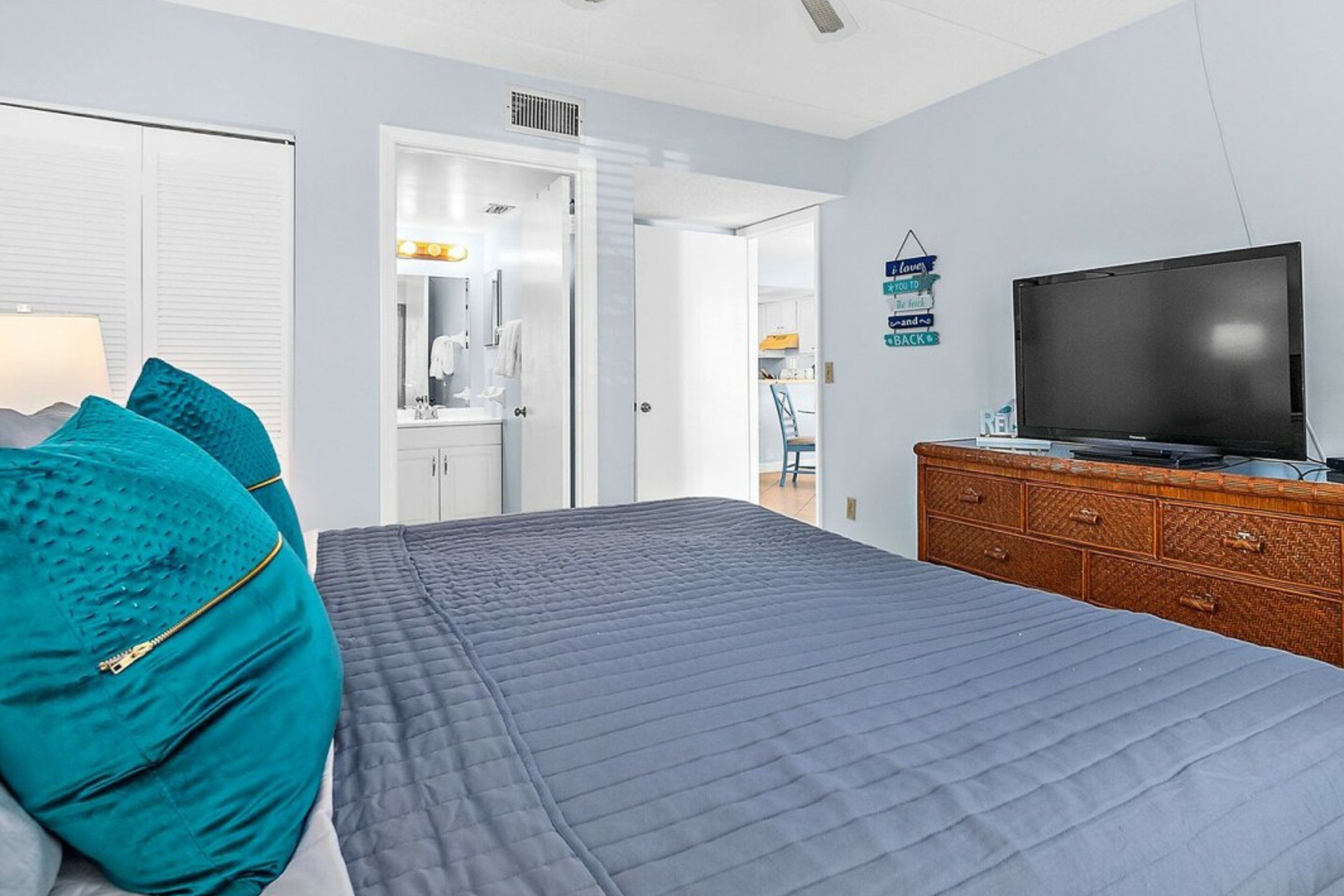 A bedroom with a bed, a dresser, a television, and a bathroom visible through an open door.
