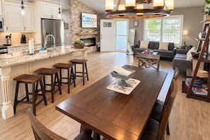 Een ruime keuken met een centraal eiland, een eetgedeelte met een lange tafel en een woonkamer met een bank en een open haard.