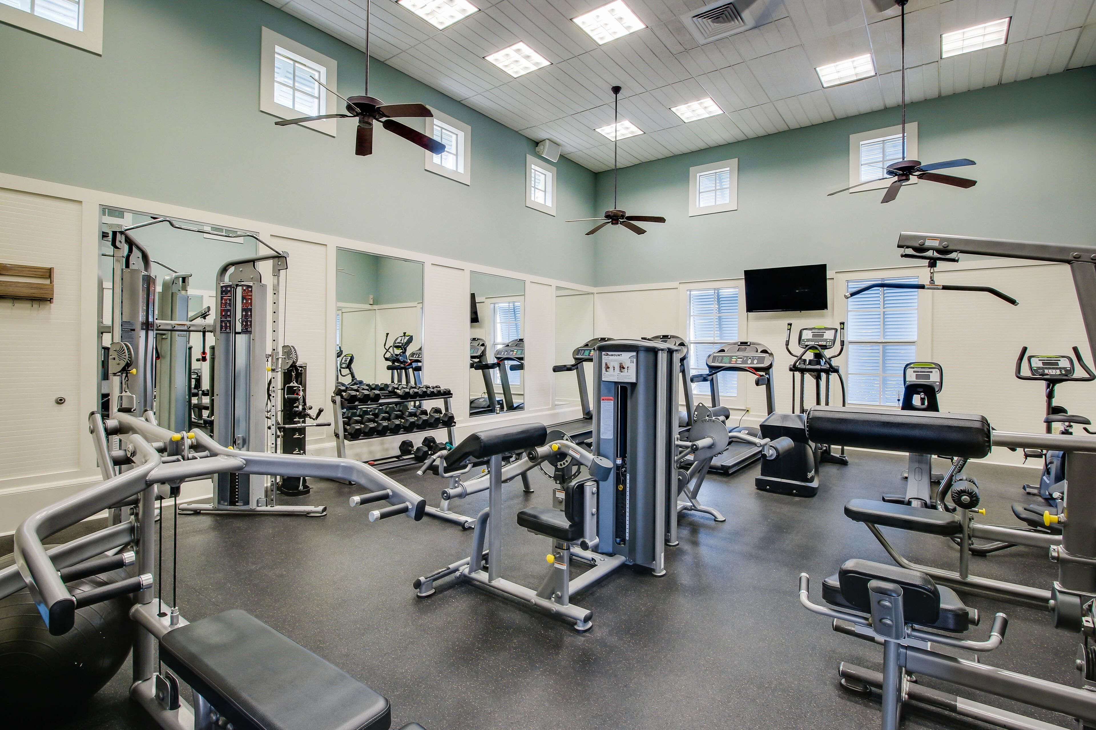 A gym with various exercise machines, mirrors, and ceiling fans.