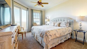 A hotel room with a large bed, a desk, a chair, a ceiling fan, a lamp, and a view of the outdoors. - Oceanfront Condo With Great Ocean View, Balcony, Fast Wifi & 2 Shared Pools (Fernandina Beach)