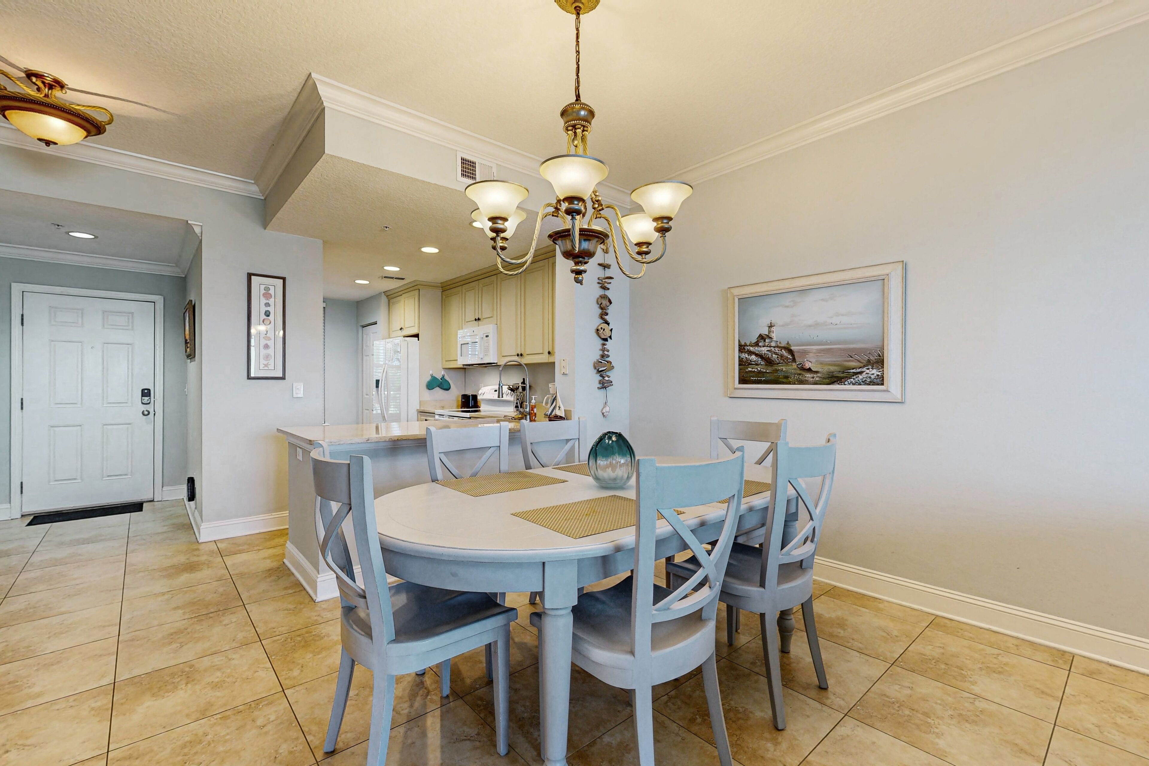 A dining area with a round table, four chairs, a chandelier, and a kitchen in the background.