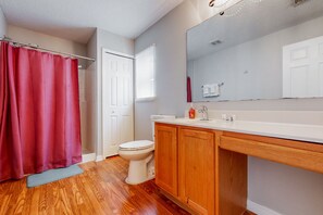 Une salle de bain avec un plancher en bois, des toilettes blanches, un meuble-lavabo en bois avec un miroir et un rideau de douche rouge.
