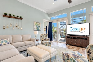A spacious living room with a sofa, ottoman, and a television displaying 'Casago Vacation Rentals'. - Palm-surrounded Stunner With Enclosed Pool & Water Views - Dog-friendly (Palm Coast)