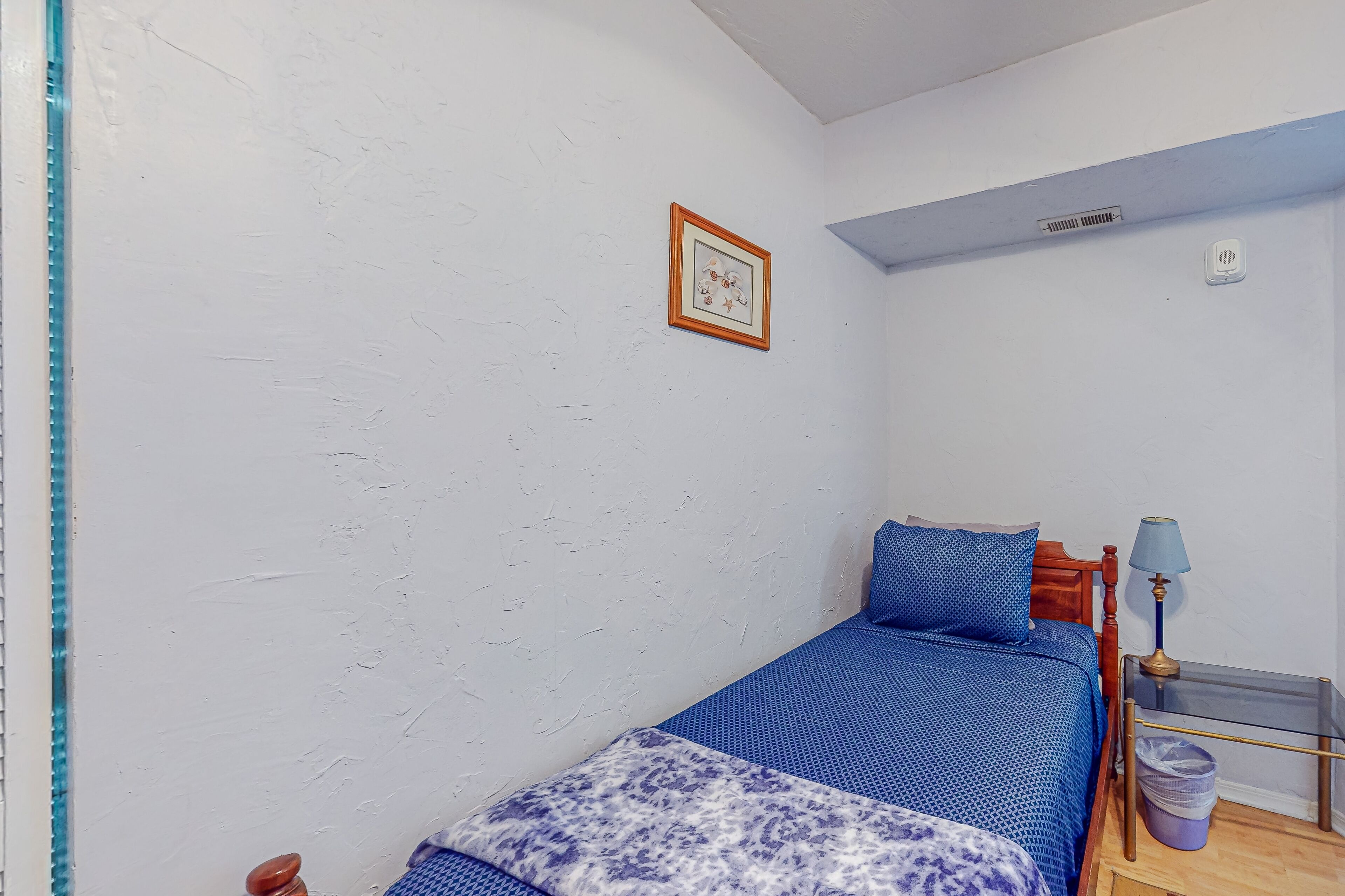 Une petite chambre avec un lit simple, une couvre-lit bleue, une tête de lit en bois, une petite table avec une lampe et un tableau accroché au mur.