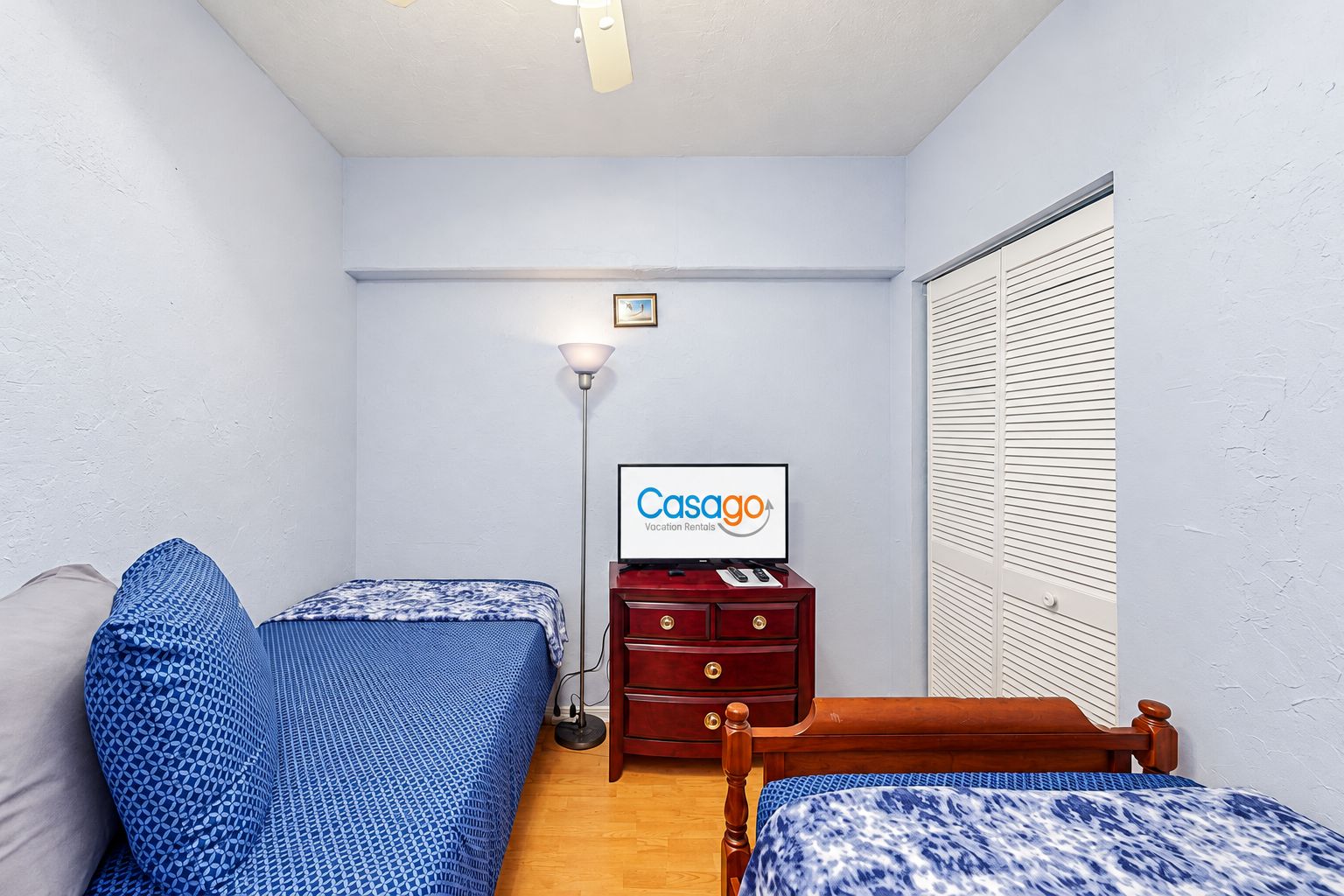 A small, well-lit room with two beds, a nightstand, a desk with a computer, and a window with blinds.