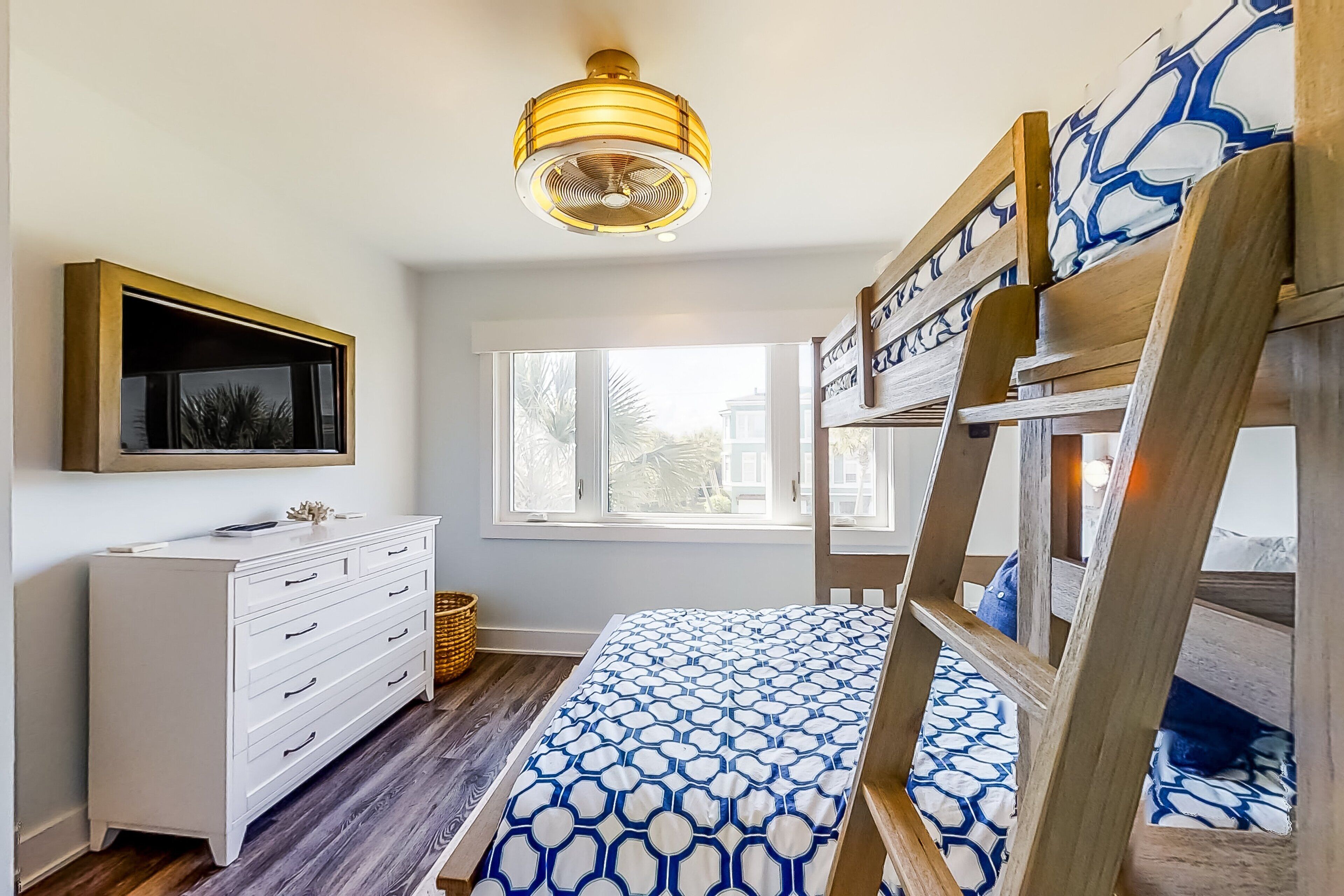 A bedroom with a bunk bed, a dresser, a television, and a window with a view of palm trees.