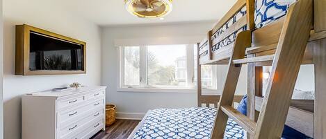 A bedroom with a bunk bed, a dresser, a television, and a window with a view of palm trees.