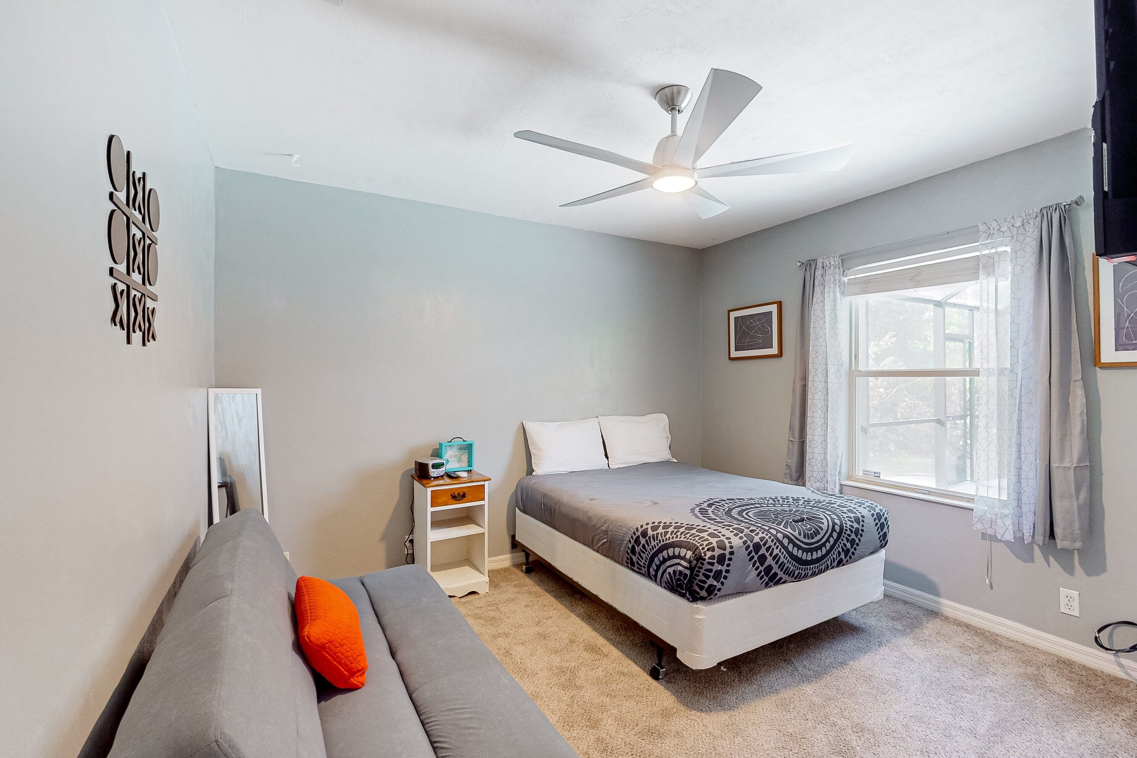 A bedroom with a bed, a sofa, a ceiling fan, a window with curtains, and a small nightstand with a lamp.