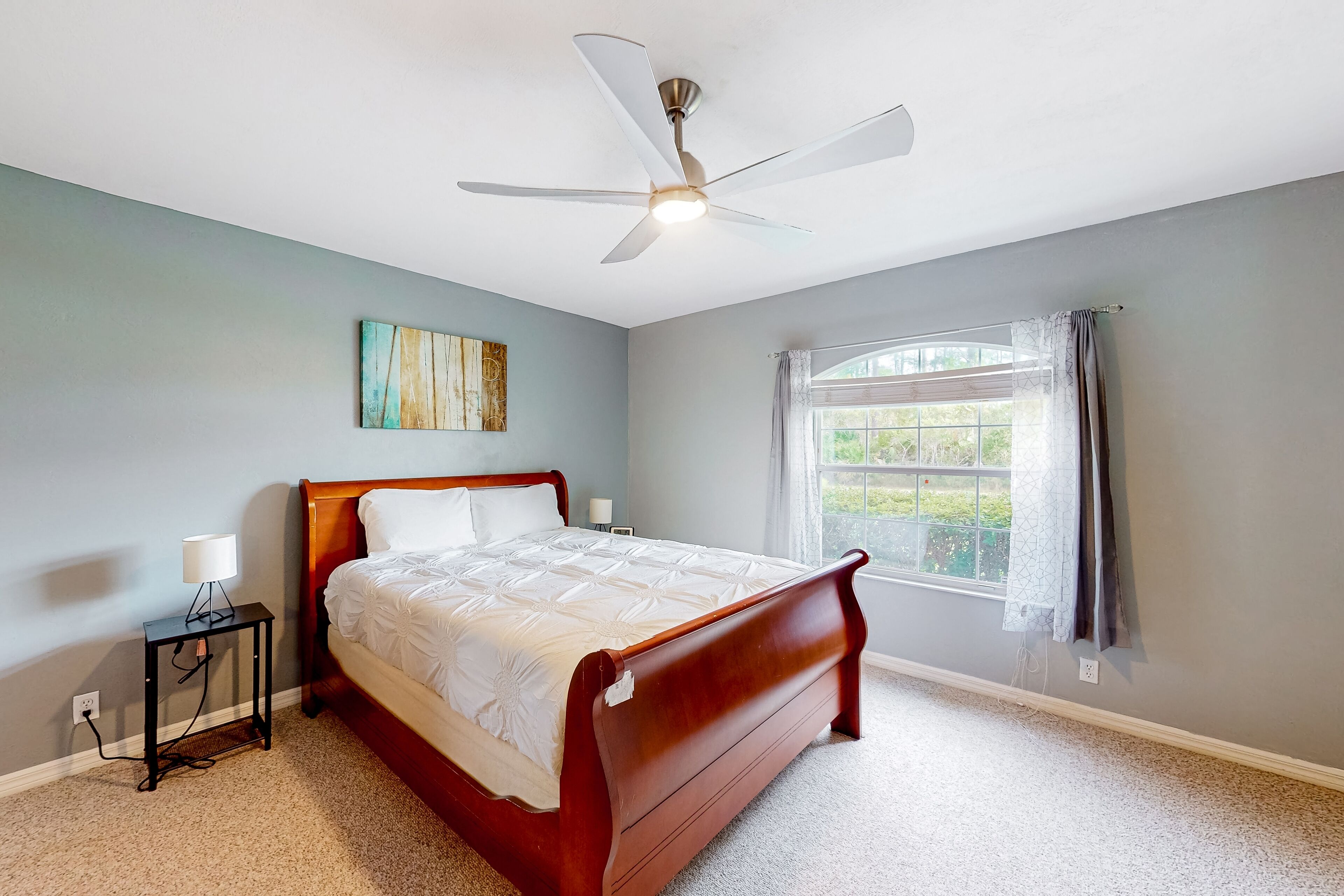Une chambre à coucher comprenant un lit, une table de chevet, un ventilateur de plafond, une fenêtre avec des rideaux et un tableau mural.