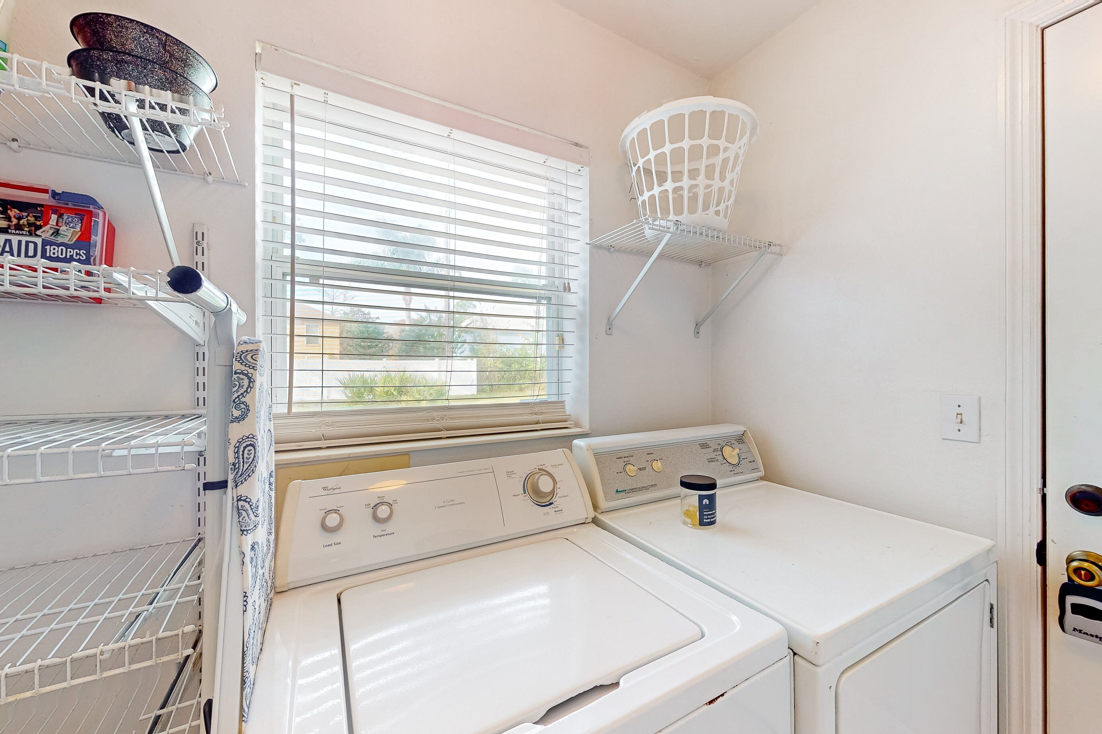 Un cuarto de lavado con dos lavadoras y secadoras apiladas, una ventana con persianas, un estante con una cesta y una caja de detergente.