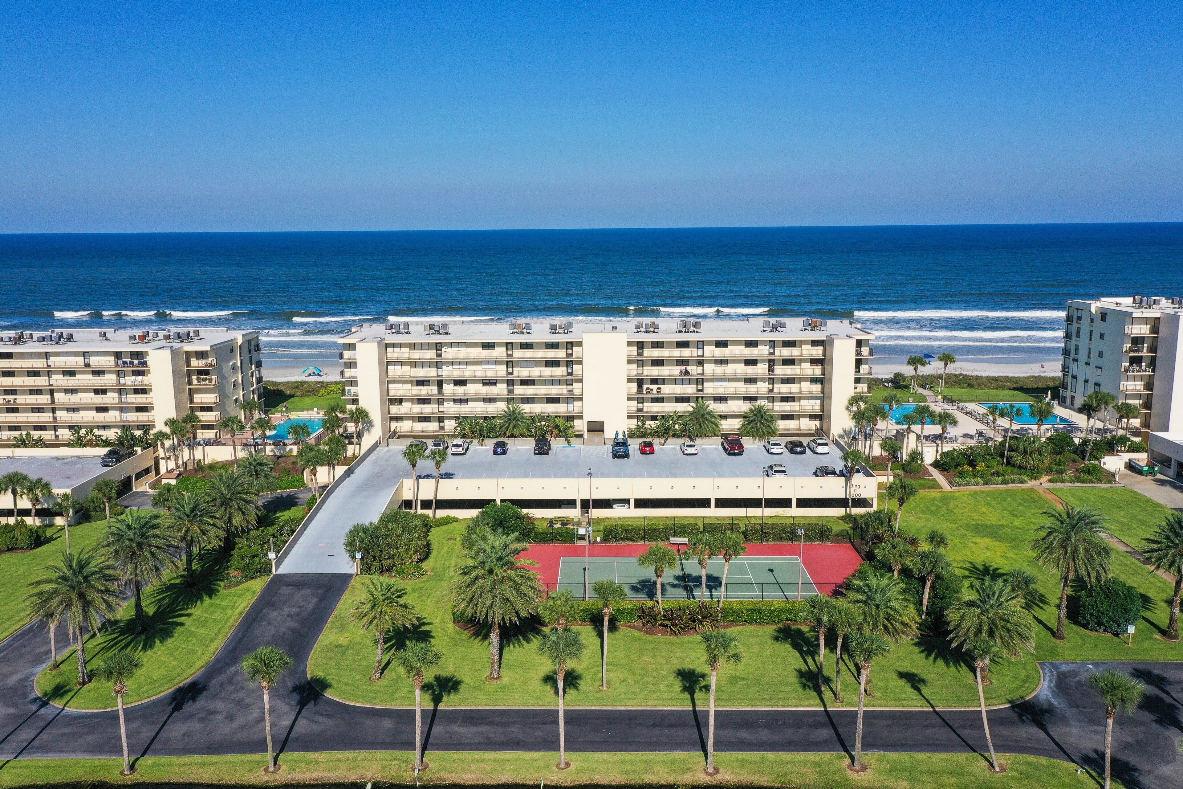 A multi-story hotel with a parking lot, a tennis court, and a view of the ocean.