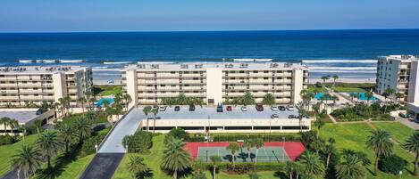A multi-story hotel with a parking lot, a tennis court, and a view of the ocean.