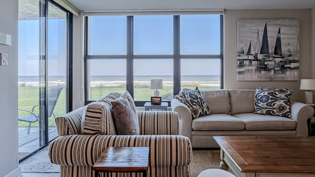 A living room with a large window, a sofa, a striped chair, and a wooden table. - Oceanfront Condo With Beach Access, Patio, Outdoor Pools, Tennis Court, W/d, AC (St. Augustine)
