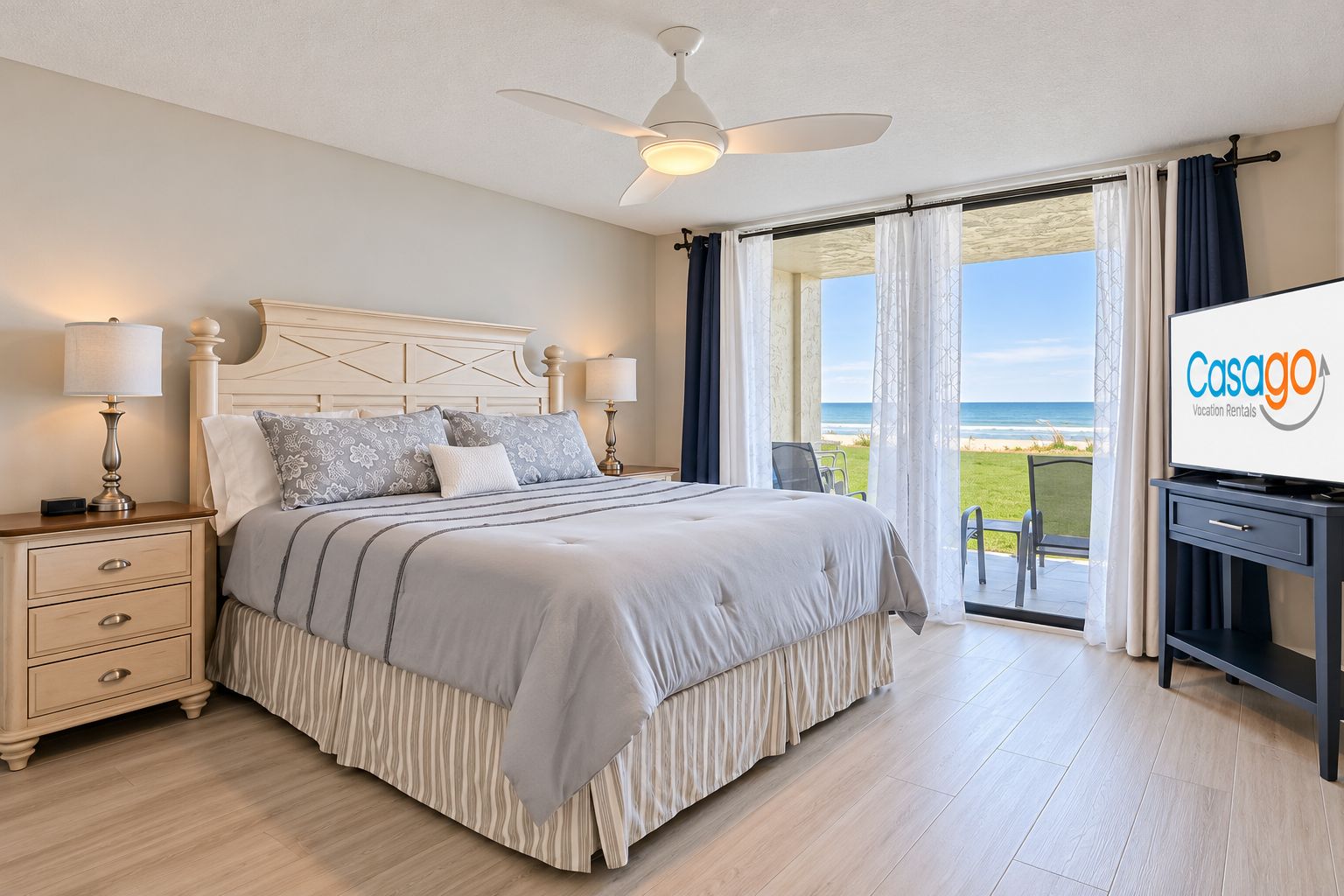 A bedroom with a bed, bedside tables, a ceiling fan, and a view of the beach through the window.