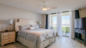 A bedroom with a bed, bedside table, lamp, TV, and a view of the outdoors. - Oceanfront Condo With Beach Access, Patio, Outdoor Pools, Tennis Court, W/d, AC (St. Augustine)