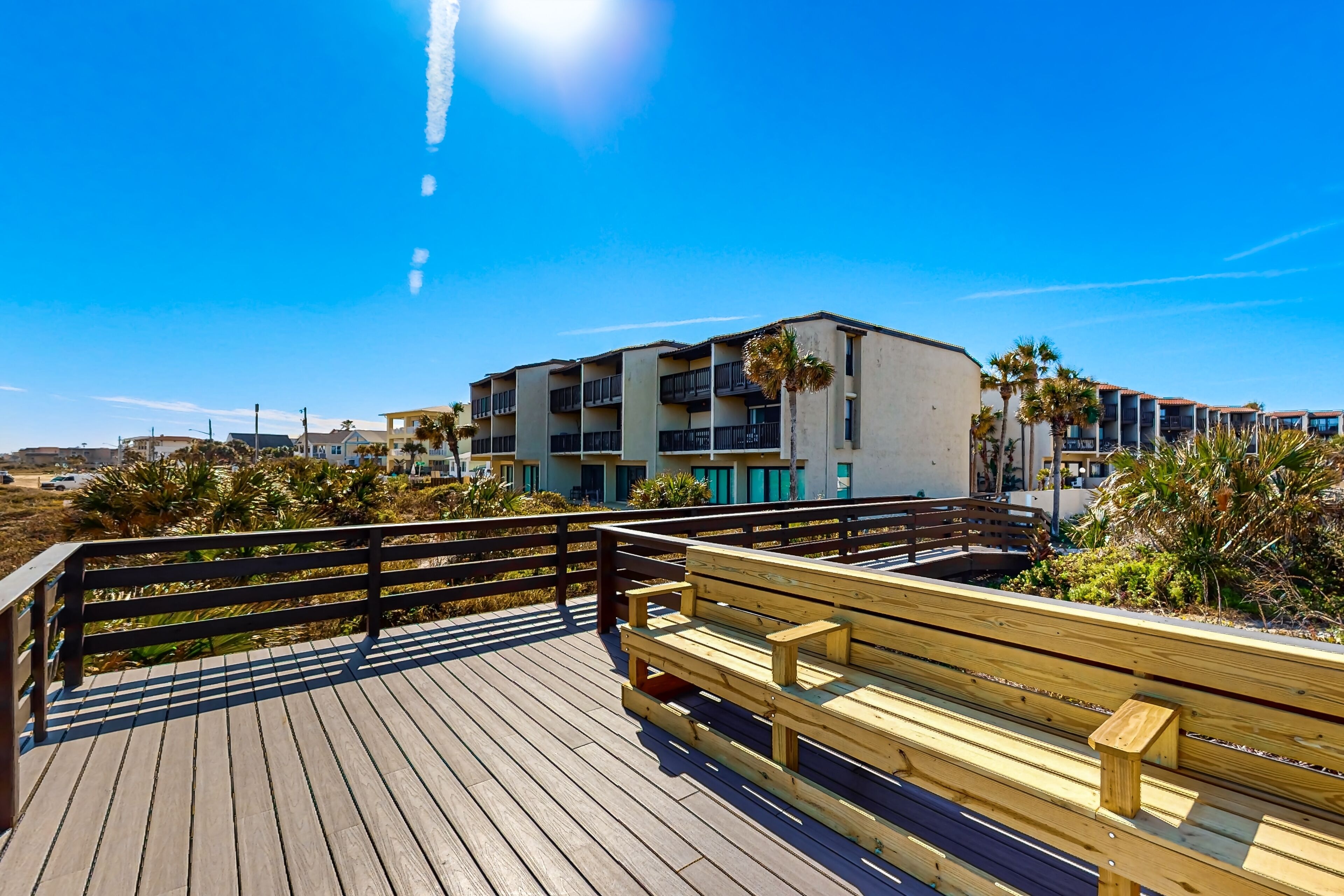 A wooden boardwalk with a bench, overlooking a beachfront with buildings and palm trees.