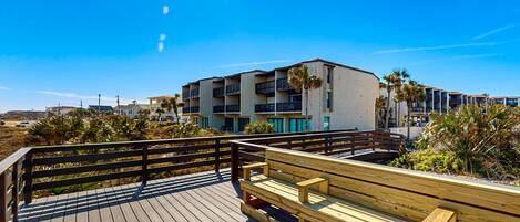 A wooden boardwalk with a bench, overlooking a beachfront with buildings and palm trees.