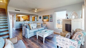 Ein geräumiges Wohnzimmer mit einem hellblauen Sofa, einem weißen Couchtisch und gemusterten Sesseln. Im Hintergrund ist eine Küche zu sehen, und eine Treppe führt auf eine obere Ebene.