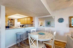 A kitchen with wooden cabinets, a dining area with a round table and white chairs, and a small stool. - Oceanfront Condo With Resort Amenities, Beach Access and Pool (St. Augustine)
