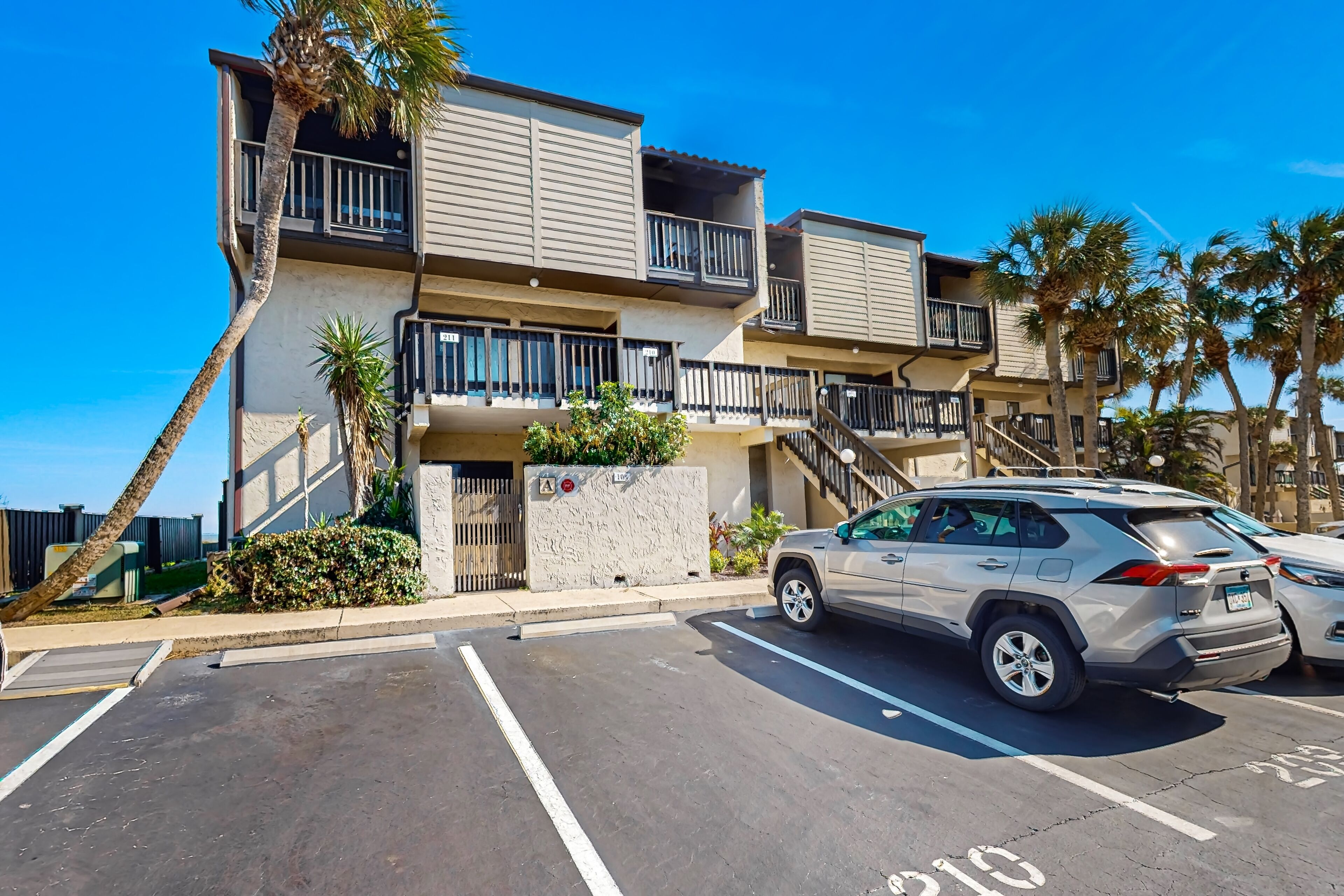 A multi-story residential building with balconies, a parking lot, and palm trees.