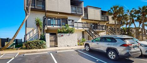 A multi-story residential building with balconies, a parking lot, and palm trees.