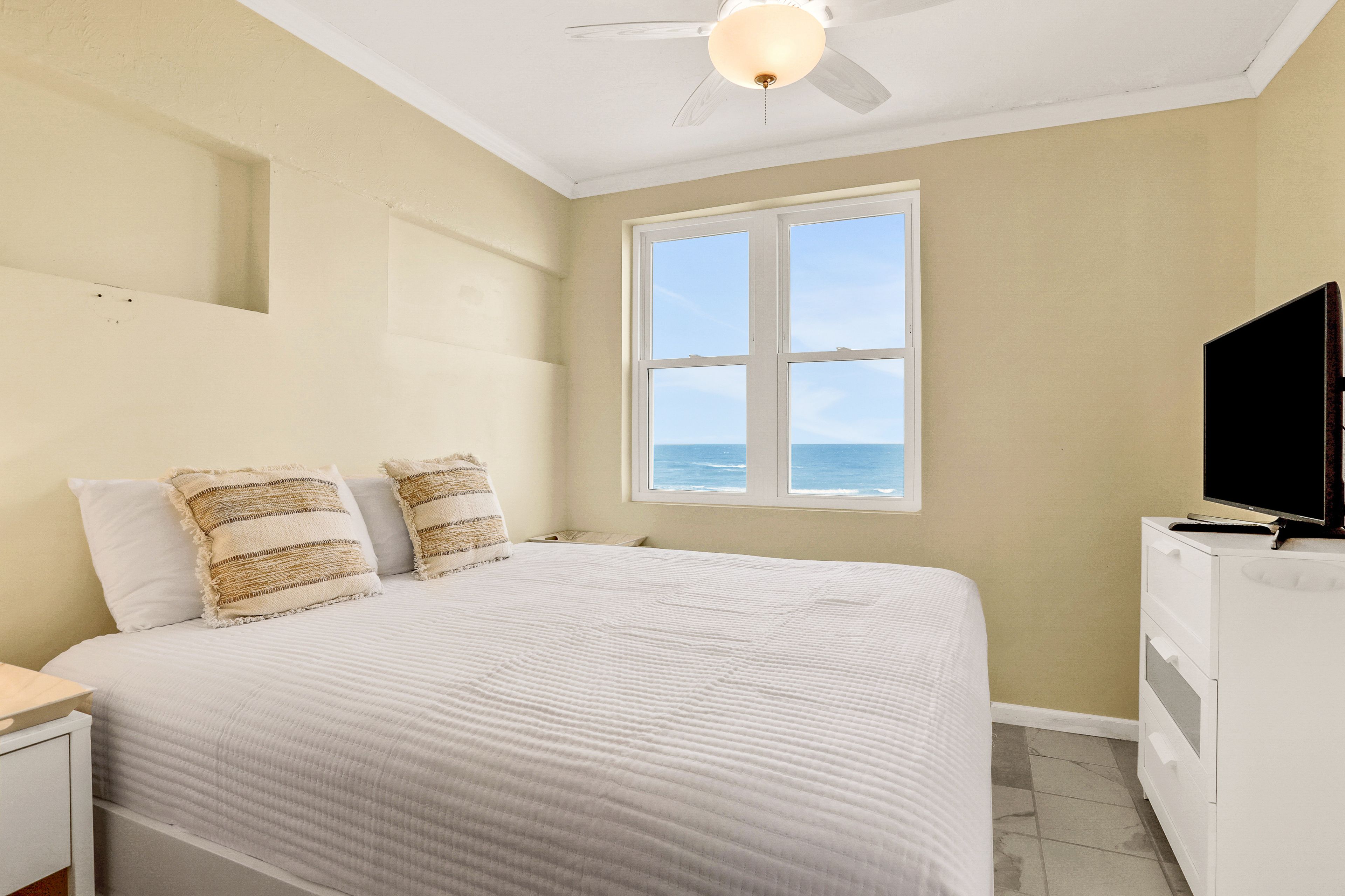 Une chambre à coucher avec un lit, un téléviseur posé sur une commode, un ventilateur de plafond et une fenêtre donnant sur l’océan.