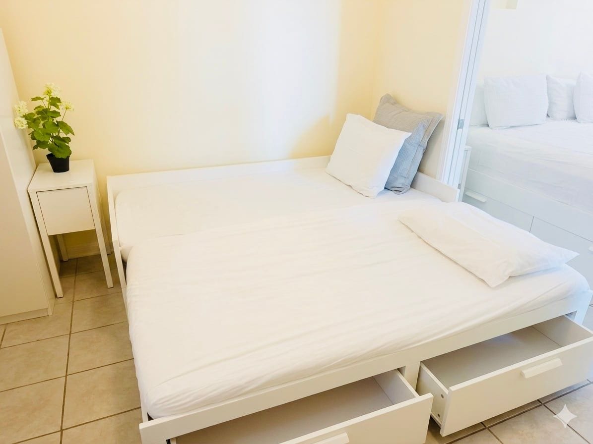Una cama con colchón y almohadas blancas, una mesita de noche con una planta y un cajón debajo de la cama.