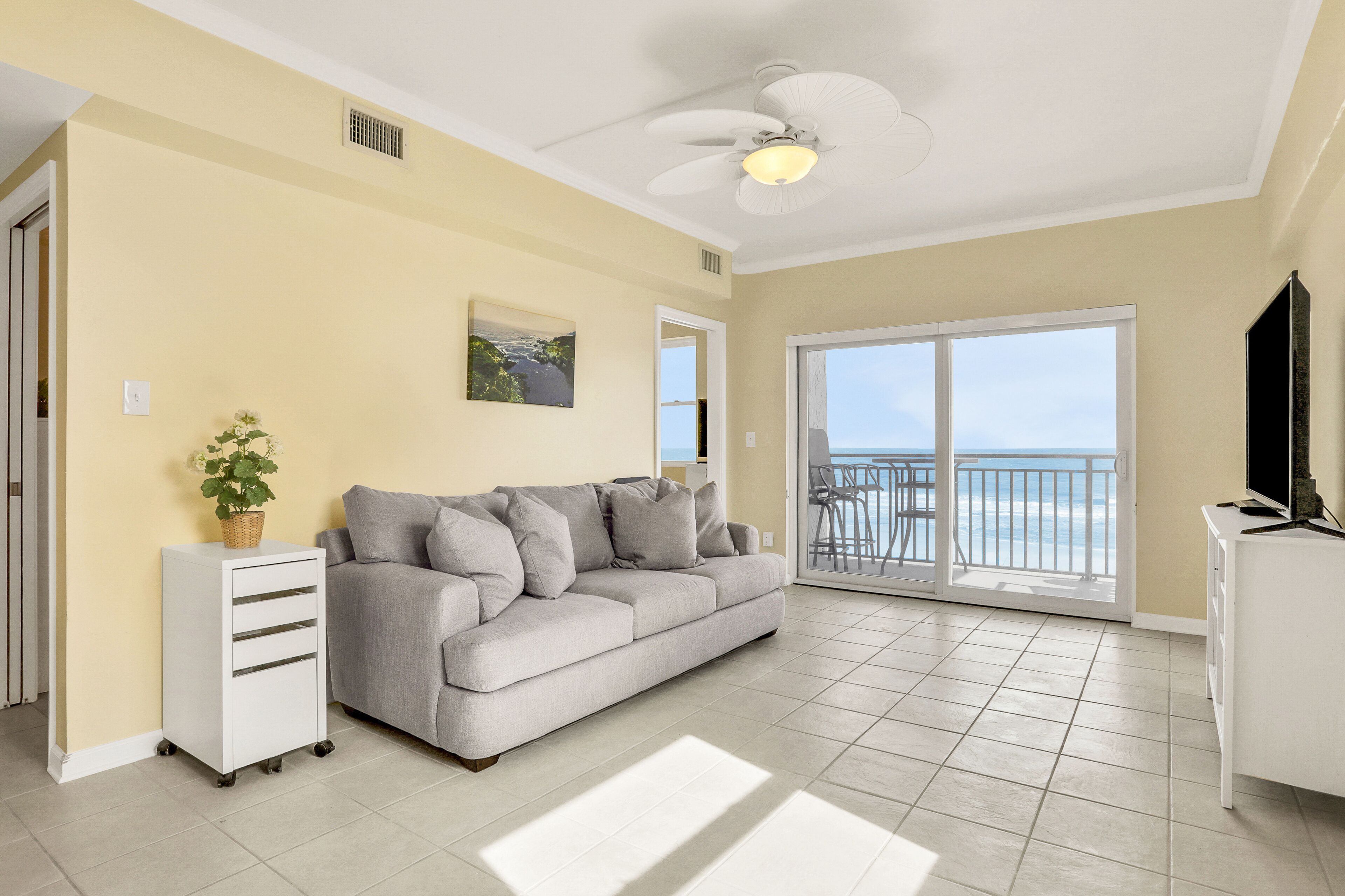 A living room with a sofa, a small white cabinet, a ceiling fan, a large window with a view, and a balcony.