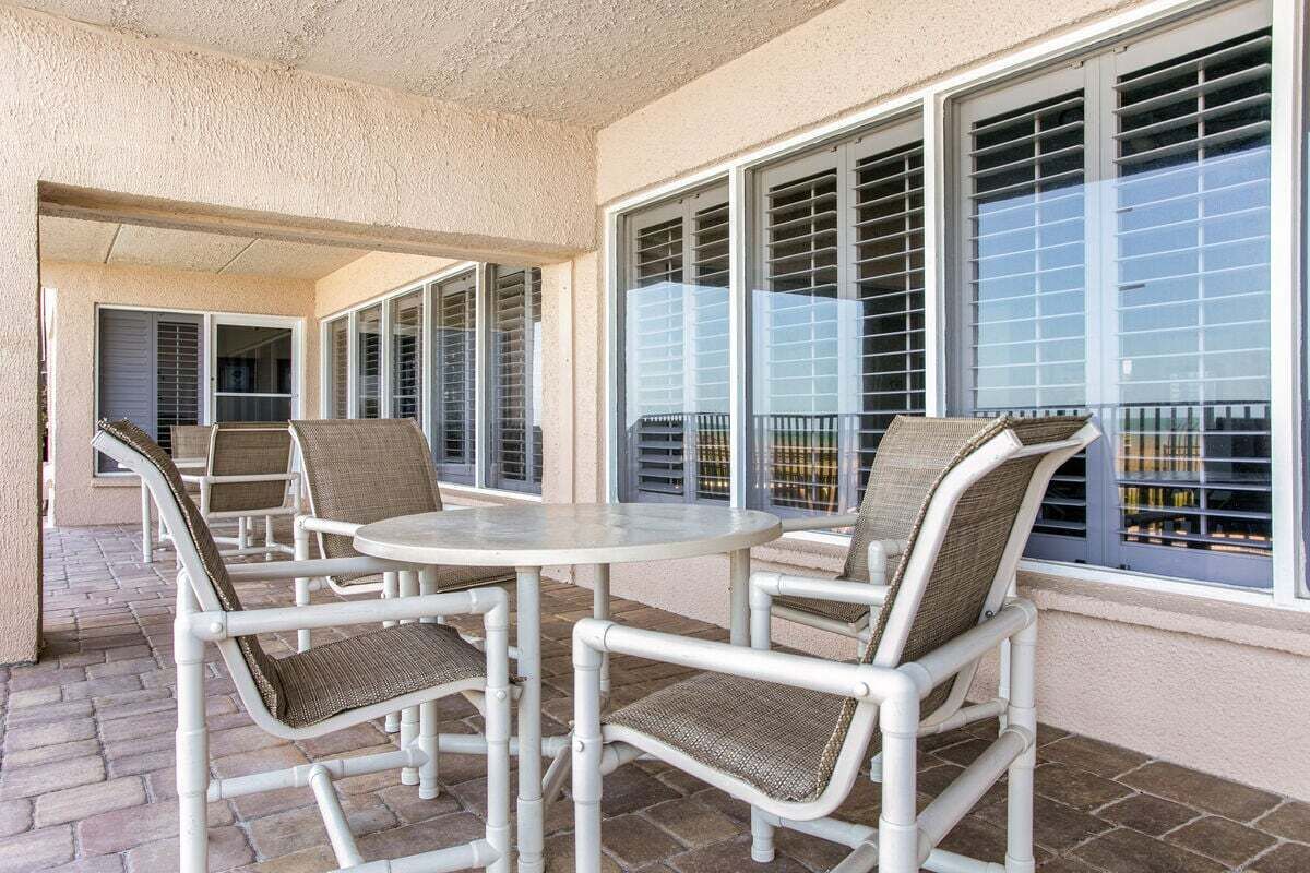 Un espace extérieur aménagé en terrasse, avec des chaises en métal blanc et une table ronde, de grandes fenêtres équipées de stores et une vue sur le paysage urbain.