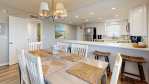 A modern kitchen with white cabinets, a central island, and a dining area with a wooden table and white chairs. - Oceanfront Corner Condo With Sunrise-view Balcony, Pool & 1-minute Walk to Beach (Fernandina Beach)