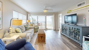 A spacious living room with a sofa, a coffee table, a TV on a cabinet, and a view to the outside. - Oceanfront Corner Condo With Sunrise-view Balcony, Pool & 1-minute Walk to Beach (Fernandina Beach)