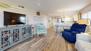 A living room with a television, a blue armchair, a white sofa, and a dining area with a white table and chairs. - Oceanfront Corner Condo With Sunrise-view Balcony, Pool & 1-minute Walk to Beach (Fernandina Beach)