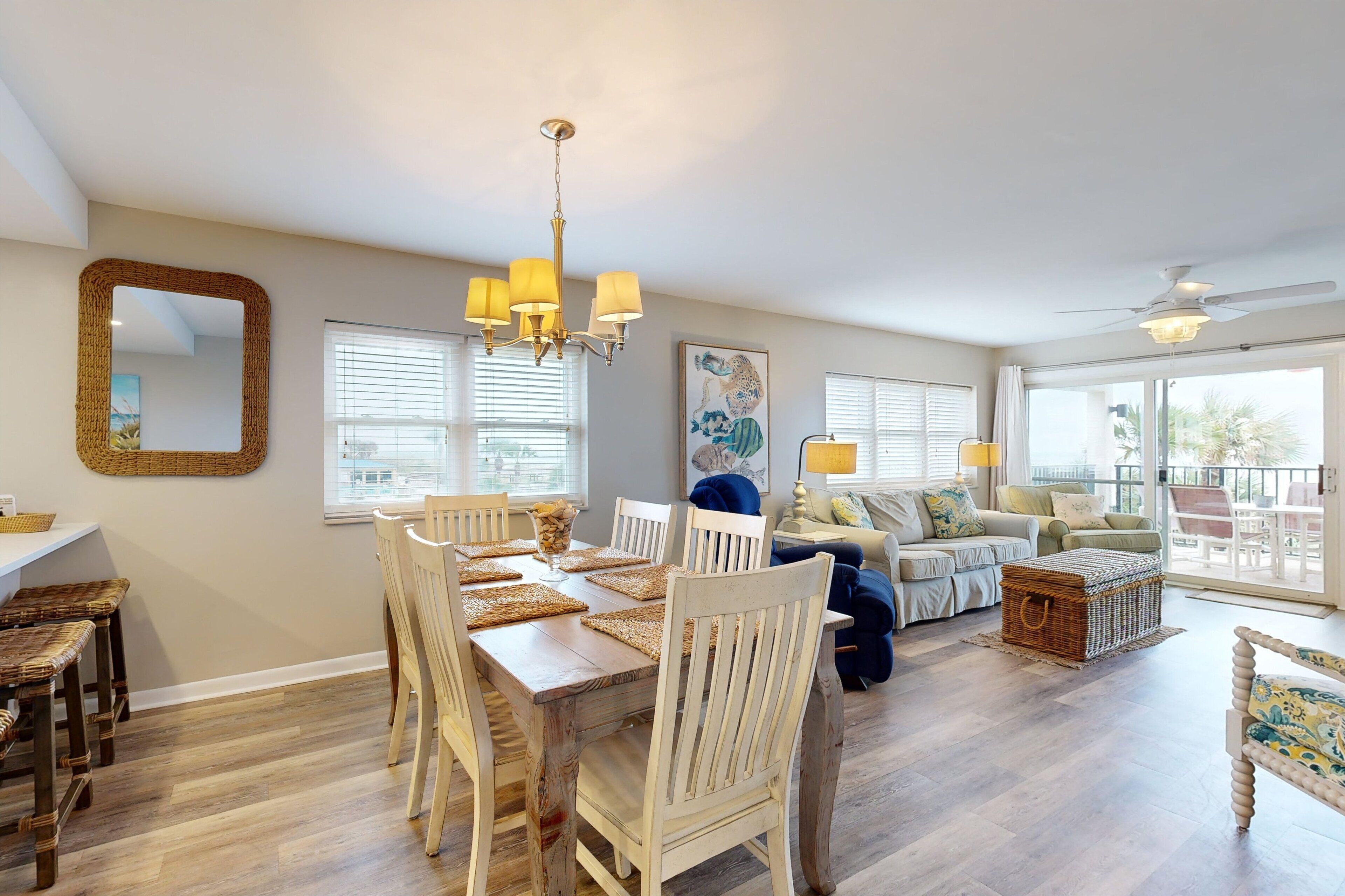 A spacious living area with a dining table, chairs, and a large mirror. There is a chandelier, a ceiling fan, and a view through large windows.