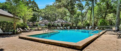 Una piscina rectangular con borde de ladrillo, rodeada de sillas de descanso y árboles.