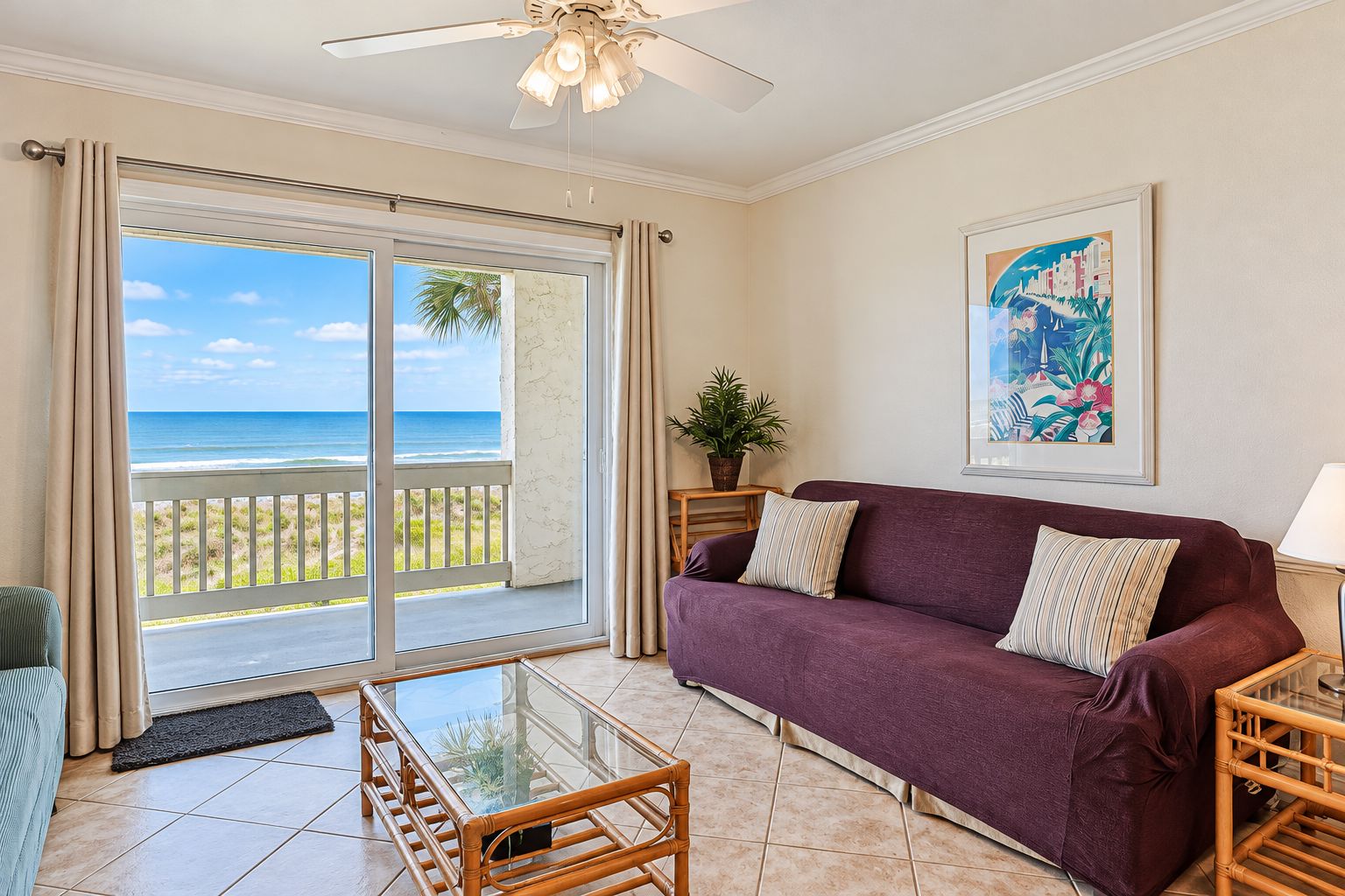 A room with a glass door leading to a balcony with a view of the ocean, a purple sofa, a wooden coffee table, and a framed picture on the wall.