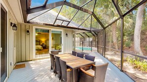 Une terrasse couverte, avec une table en bois et des chaises en osier, donnant sur une piscine et une végétation luxuriante.