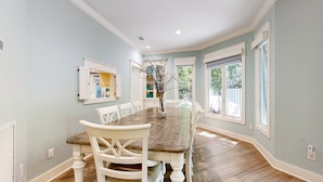 Une salle à manger avec une table en bois et des chaises blanches, de grandes fenêtres et une vue sur l’extérieur.
