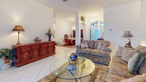 A spacious living room with a glass coffee table, a patterned sofa, and a wooden cabinet with decorative items. - Prime House With Lanai, Private Pool, Bbq, and Water Views (Palm Coast)