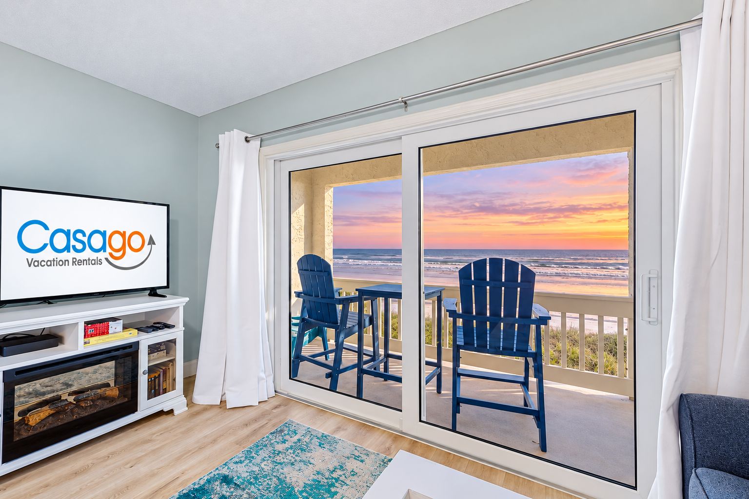 A room with a television displaying 'Casago Vacation Rentals,' a balcony with two chairs, and a view of the ocean at sunset.