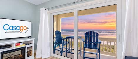 Una habitación con una televisión mostrando 'Casago Vacation Rentals', un balcón con dos sillas y vista al océano al atardecer.