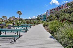 A pathway lined with green picnic tables and benches, leading to a building with balconies and a view of palm trees and other greenery. - Oceanfront Condo With Large Balcony, Pool, Central AC & Views (Fernandina Beach)