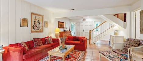 Un salon spacieux avec deux canapés rouges, une table basse en bois et un tapis à motifs. Un escalier mène à l’étage supérieur.