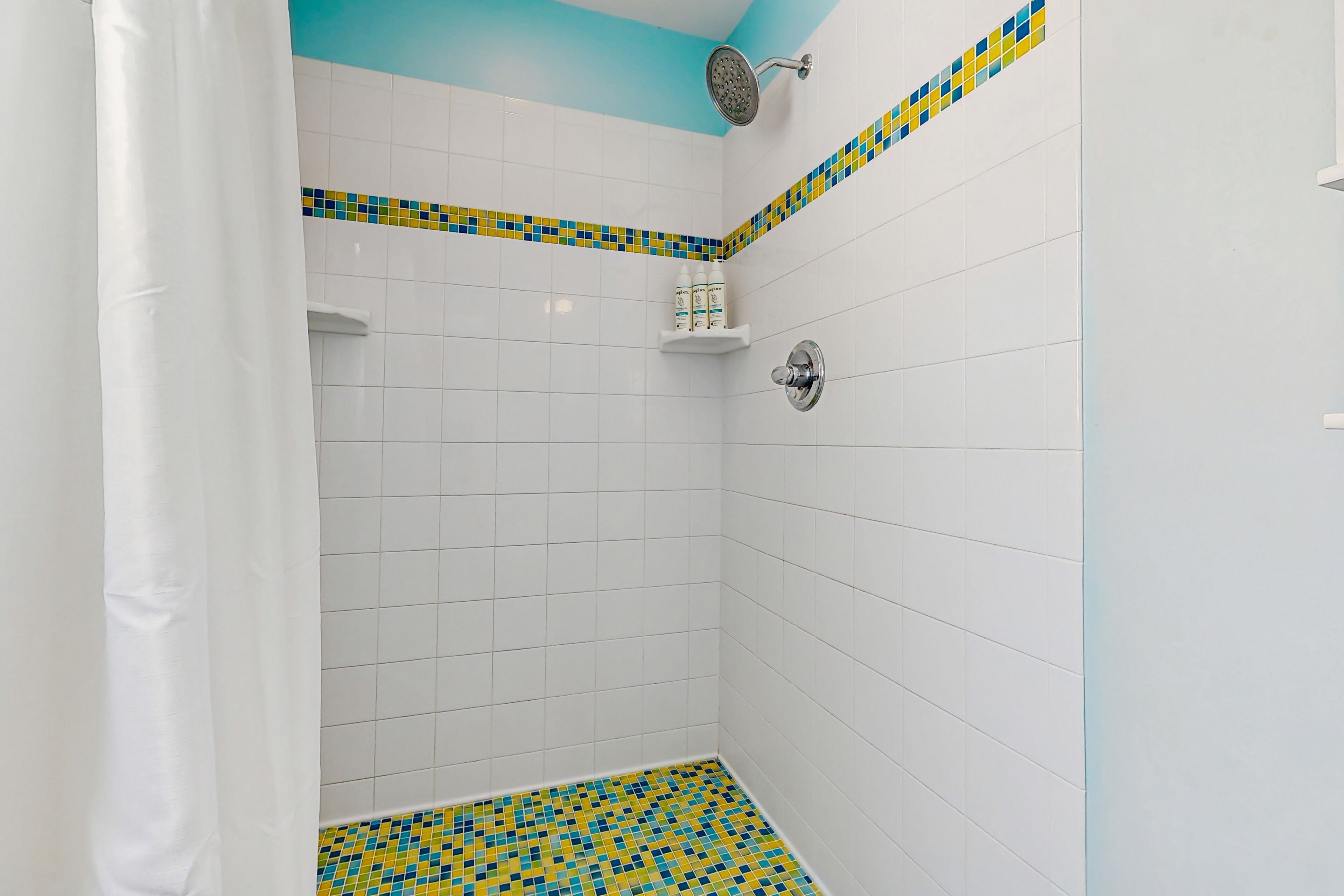 A shower area with white tiles, a blue ceiling, and a yellow and blue mosaic border.