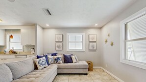 A living room with a sectional sofa, a kitchen in the background, and framed artwork on the walls. - Spacious Beach House With Private Pool, Firepit and Enclosed Yard (St. Augustine)