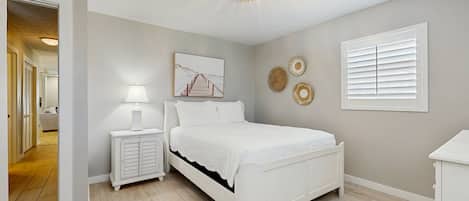 A bedroom with a white bed, a nightstand with a lamp, a ceiling light fixture, and a window with blinds.