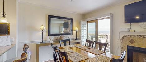 A dining area with a wooden table, chairs, and placemats. There is a fireplace, a television, and a window with shutters.