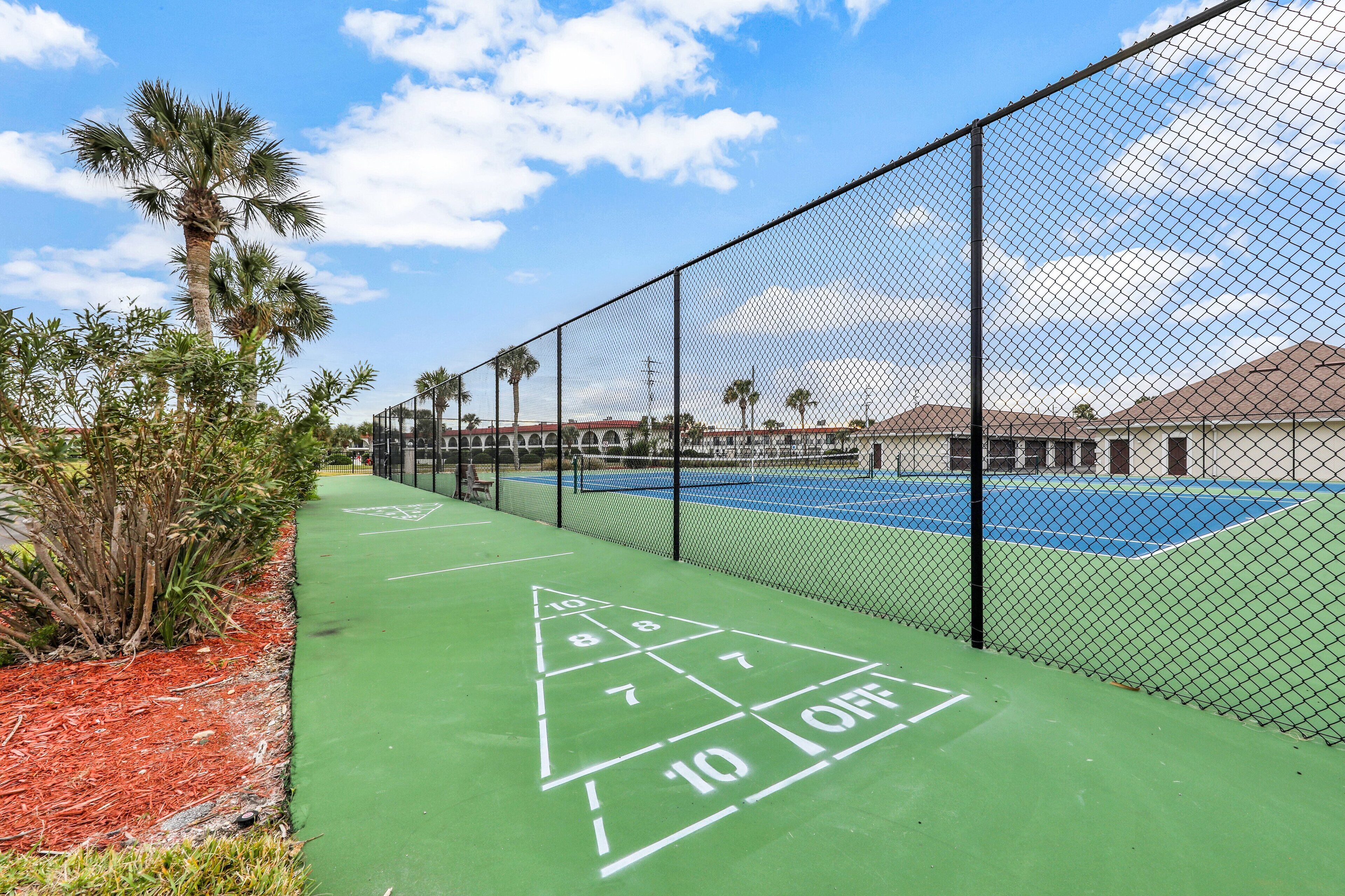 Un court de tennis avec une zone verte aménagée pour le jeu de palets, entouré d’une clôture en treillis métallique et de palmiers.