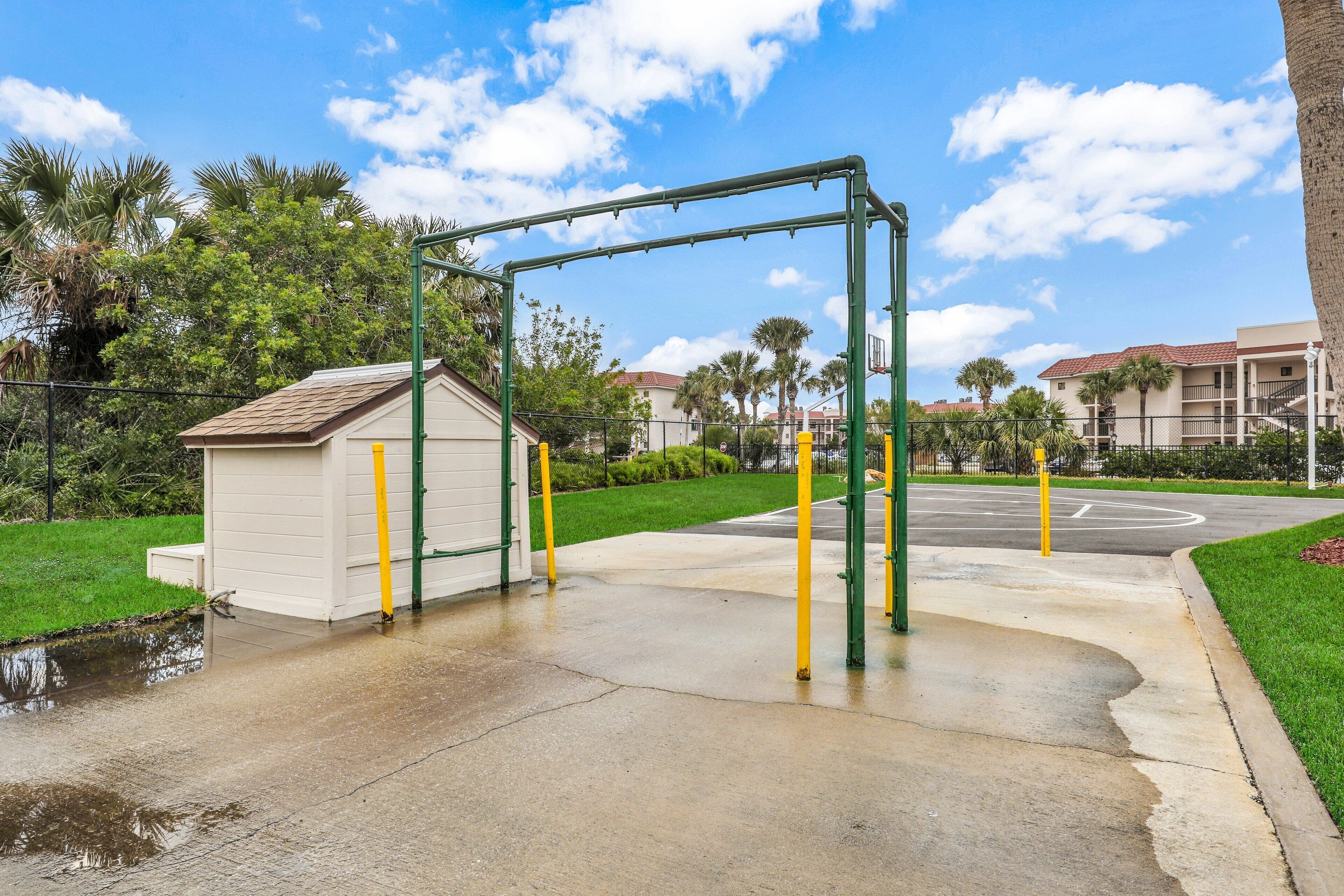 A playground with a swing set and a small shed, surrounded by a fence and palm trees.