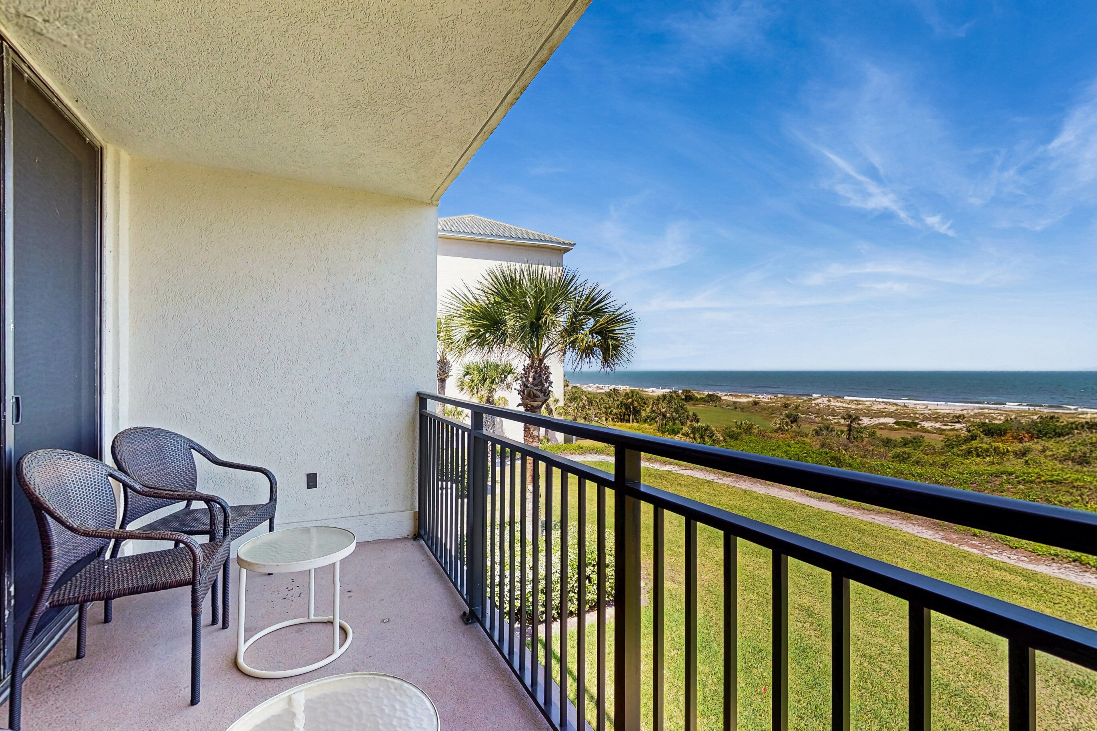 A balcony with two wicker chairs and a small table, overlooking a grassy area and the ocean.